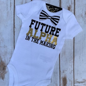 May include: White baby onesie with black and white striped bow tie and gold lettering that reads "Future Alpha in the Making".