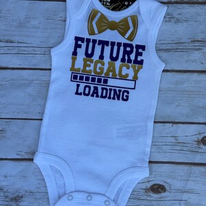 May include: White baby bodysuit with a gold bow tie graphic and the words "FUTURE LEGACY LOADING" in purple and gold. The bodysuit has a sleeveless design and snap closures at the bottom.