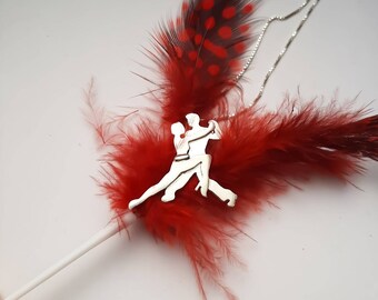 May include: A silver necklace with a pendant featuring a silhouette of two people dancing the tango. The necklace is displayed on a red feather boa.