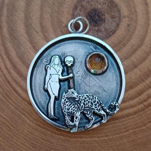 Puede incluir: Colgante de plata con un dise&ntilde;o detallado. Presenta una mujer, una calavera y un leopardo. Una piedra preciosa amarilla est&aacute; engastada en el colgante. El colgante es redondo con un lazo en la parte superior para una cadena.