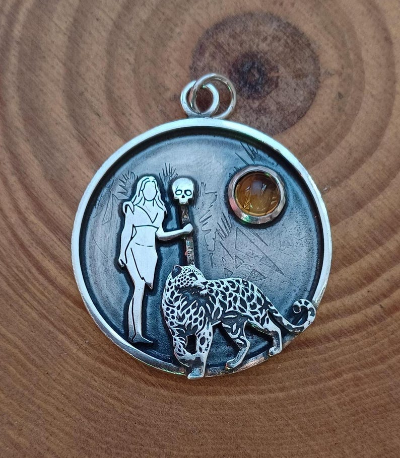 Puede incluir: Colgante de plata con un dise&ntilde;o detallado. Presenta una mujer, una calavera y un leopardo. Una piedra preciosa amarilla est&aacute; engastada en el colgante. El colgante es redondo con un lazo en la parte superior para una cadena.