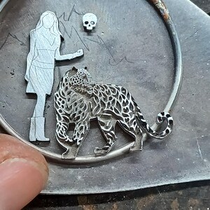 Puede incluir: Colgante de metal plateado con un dise&ntilde;o de mujer, leopardo y calavera. El leopardo est&aacute; intrincadamente detallado. El colgante est&aacute; sobre una superficie texturizada, con un marco circular.