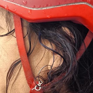 Peut inclure: Gros plan sur un casque de vélo rouge avec des gouttelettes d'eau. Le casque a une sangle rouge et un fermoir argenté. Des cheveux foncés sont visibles sous le casque.