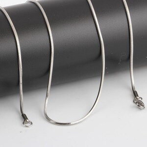 Puede incluir: Un collar de cadena de serpiente de color plateado. La cadena est&aacute; hecha de peque&ntilde;os eslabones met&aacute;licos entrelazados, creando una textura suave y flexible. El collar tiene un dise&ntilde;o sencillo con un cierre en un extremo y un lazo en el otro.