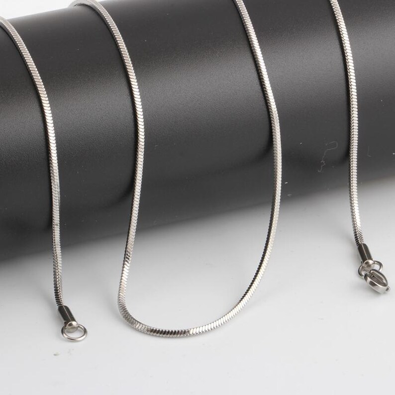 Puede incluir: Un collar de cadena de serpiente de color plateado. La cadena est&aacute; hecha de peque&ntilde;os eslabones met&aacute;licos entrelazados, creando una textura suave y flexible. El collar tiene un dise&ntilde;o sencillo con un cierre en un extremo y un lazo en el otro.