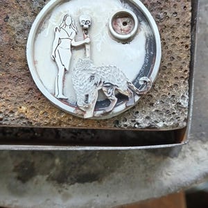 Puede incluir: Colgante de plata con dise&ntilde;o circular. El colgante presenta una mujer, una calavera y una criatura parecida a un lobo. El colgante tiene un lazo en la parte superior para una cadena. El fondo es una superficie marr&oacute;n texturizada.