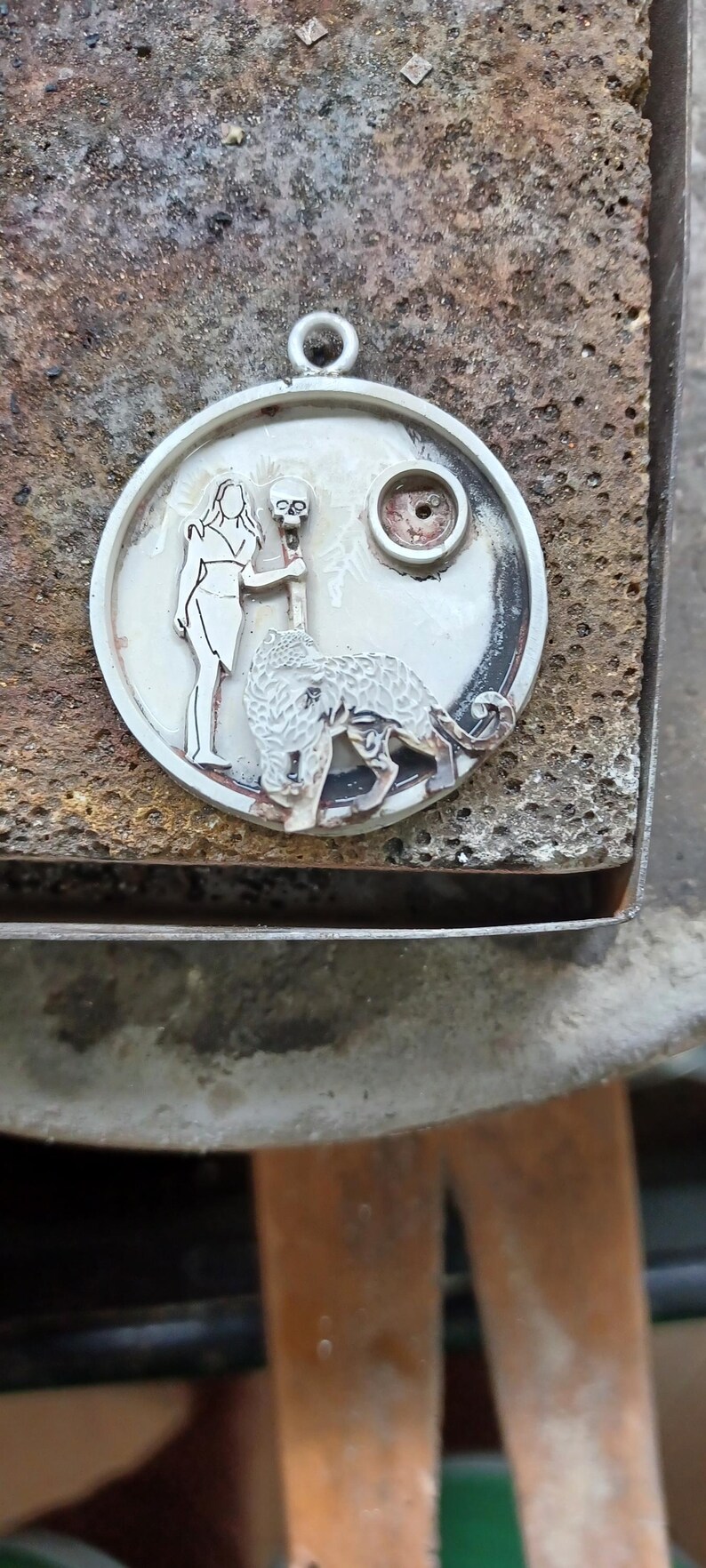 Puede incluir: Colgante de plata con dise&ntilde;o circular. El colgante presenta una mujer, una calavera y una criatura parecida a un lobo. El colgante tiene un lazo en la parte superior para una cadena. El fondo es una superficie marr&oacute;n texturizada.