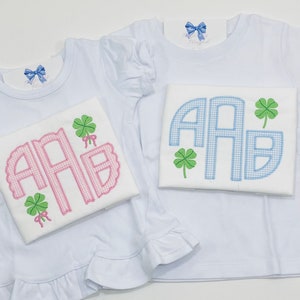 May include: Two white short-sleeved shirts with ruffled hems. One shirt has a pink monogram with a shamrock and bow. The other shirt has a blue monogram with a shamrock.