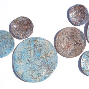 Peut inclure: Un ensemble de huit disques muraux en céramique circulaire avec une finition texturée. Les disques présentent un mélange de tons bleus, bruns et argentés métalliques tachetés avec un bord organique irrégulier, disposés en groupe.