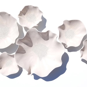 May include: A collection of seven white ceramic wall sculptures shaped like ruffled flowers. Each piece features a smooth, curved outer edge and a textured, granular center. The sculptures vary in size and are arranged in a cluster.
