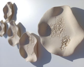 May include: A collection of five off-white ceramic wall sculptures with organic, wavy edges and textured, pebbled centers. Each piece features a matte finish and irregular, undulating forms that create depth through light and shadow.
