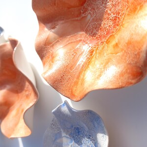 May include: Close-up of ceramic wall art in shades of orange, white, and blue. The pieces have a wavy, organic shape, with a glossy finish. The art is likely for home decor.