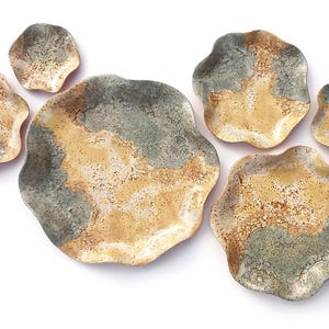 Puede incluir: Un conjunto de siete platos de cerámica de forma irregular. Los platos presentan un diseño moteado en tonos grises, beige y crema. El plato más grande mide unos 30 cm de diámetro. Un juego de vajilla decorativo.