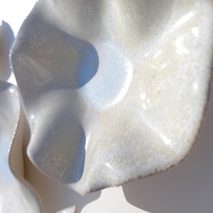 May include: Close-up of three white, organically shaped ceramic wall art pieces. The pieces have a glossy, textured surface with subtle color variations. The forms have a rippled, petal-like design, creating a sense of movement and depth.
