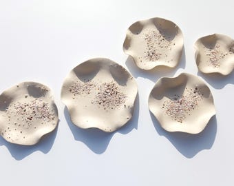 May include: Five off-white ceramic dishes with wavy, organic edges. Each dish features a textured center embedded with small, multicolored sand and shell fragments, creating a speckled appearance across the smooth, matte surface of the clay.