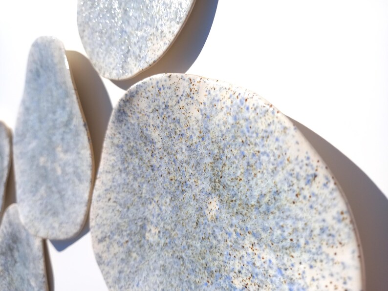 May include: A close-up of several irregularly shaped, ceramic wall art pieces. The pieces have a speckled, light blue and white glaze with brown flecks. The shapes are organic and appear to be mounted on a white wall.