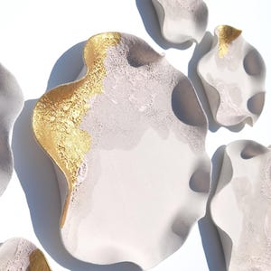 May include: A collection of five irregular, wavy-edged dishes made of a light gray, textured material. Each dish features a smooth, matte surface with a distinct section of metallic gold leaf applied to one side, creating a contrast in texture.