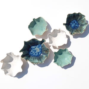 May include: An overhead shot displays seven ceramic leaf-shaped bowls in shades of white, teal, and deep blue. The bowls have a textured, organic design, with some featuring a contrasting blue interior. The arrangement is set against a bright white background.
