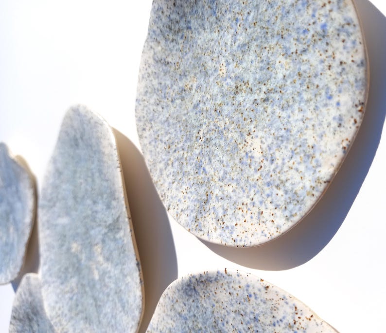 May include: A collection of irregularly shaped ceramic wall art pieces. The pieces have a speckled, light blue and white glaze with brown flecks. The organic forms are mounted on a white wall, creating a modern, minimalist aesthetic.