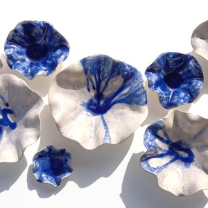 May include: A collection of handmade ceramic bowls in various sizes. The bowls are white with a blue, dripped glaze design. The bowls have a ruffled edge and are arranged on a white surface.