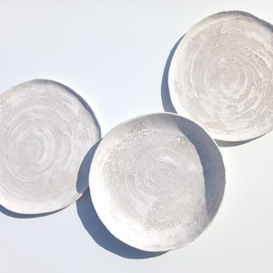 May include: Three white ceramic plates with a textured, swirling design. The plates are arranged in a triangular pattern on a white background.