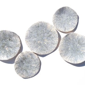 May include: Five round, ceramic plates with a speckled, light blue and white design. The plates are arranged on a white surface, with each plate's shadow visible. The plates have a unique, organic shape.
