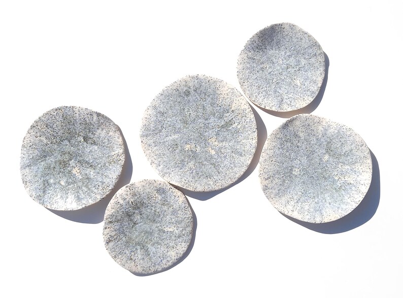 May include: Five round, ceramic plates with a speckled, light blue and white design. The plates are arranged on a white surface, with each plate's shadow visible. The plates have a unique, organic shape.