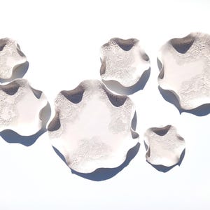 May include: A collection of five off-white ceramic wall sculptures with organic, wavy edges and a textured, cratered surface finish. Each piece features a smooth central area surrounded by a rough, porous border, creating a layered, dimensional effect.