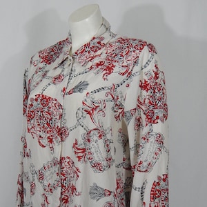 Op de afbeelding: Een wit overhemd met lange mouwen en knopen met een rode en zwarte print van kettingen, wapenschilden en bloemmotieven. Het overhemd heeft een klassieke kraag en een knoopsluiting aan de voorkant.