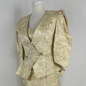 Może przedstawiać: Zestaw vintage składający się z dwóch części w jasnym złotym kolorze, z kwiatowym wzorem. Zestaw zawiera top z dekoltem w serek z przodu w stylu kopertowym i spódnicę ołówkową do kolan. Kurtka ma krótkie rękawy i ozdobne guziki.