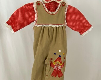 May include: A vintage baby outfit featuring a tan corduroy jumpsuit with a clown applique and colorful button accents. The outfit includes a red polka-dot long-sleeved shirt with a white lace collar and cuffs. Buttons secure the jumpsuit.