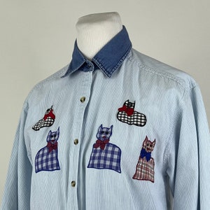 May include: A light blue denim shirt with a white and blue striped pattern. The shirt has a denim collar and features embroidered cat appliques in various colors and patterns.