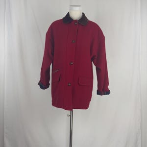 Op de afbeelding: Een rode wollen jas met een bruine kraag en knoopsluiting. De jas heeft twee voorzakken en geruite manchetten. De jas wordt gepresenteerd op een mannequin.