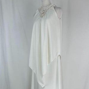 May include: A white sleeveless dress with a crochet neckline. The dress has a flowing, layered design, with a draped overlay that adds movement. The fabric appears to be lightweight, suitable for warmer weather or special occasions.