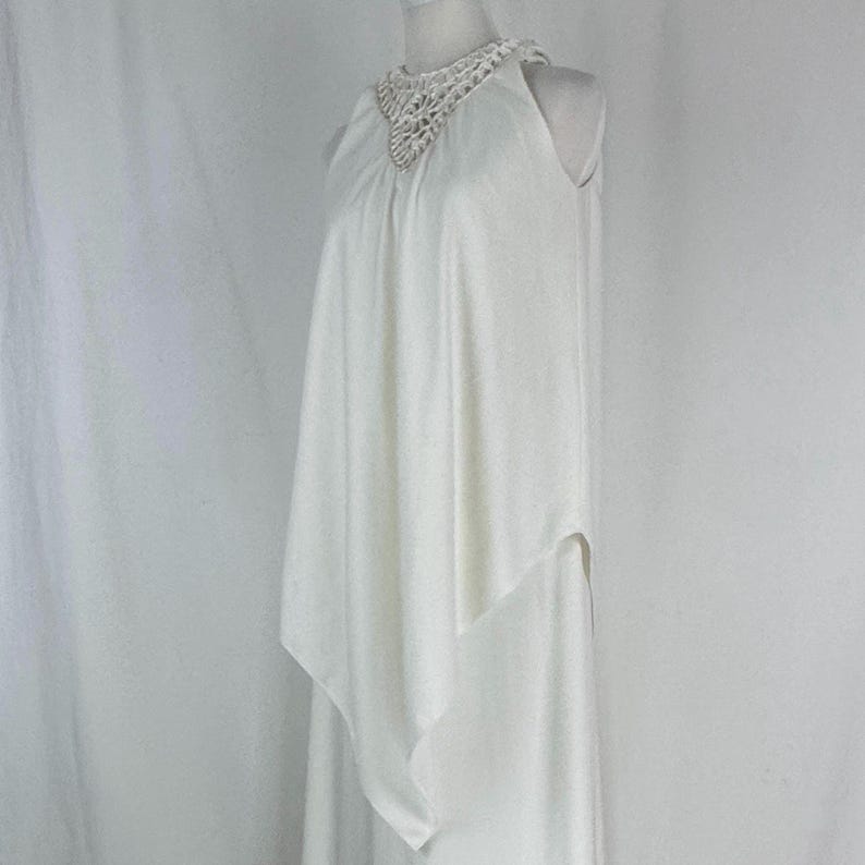 May include: A white sleeveless dress with a crochet neckline. The dress has a flowing, layered design, with a draped overlay that adds movement. The fabric appears to be lightweight, suitable for warmer weather or special occasions.