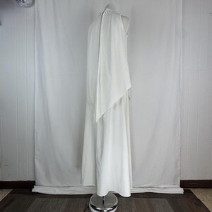 May include: A white, floor-length dress with a high neckline and a flowing cape-like overlay. The dress is displayed on a mannequin, showcasing its elegant design and silhouette. The dress appears to be made of a soft, flowing fabric.