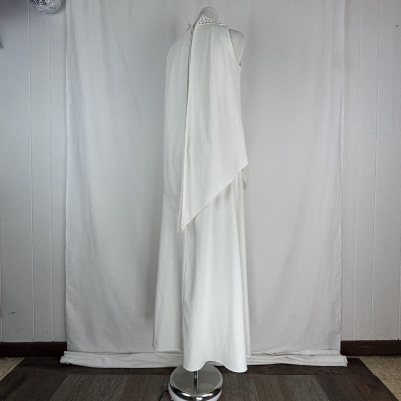 May include: A white, floor-length dress with a high neckline and a flowing cape-like overlay. The dress is displayed on a mannequin, showcasing its elegant design and silhouette. The dress appears to be made of a soft, flowing fabric.