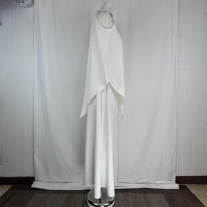May include: A white, floor-length dress with a cape overlay. The dress has a halter neckline and a flowing, layered design. The fabric appears to be a lightweight material, creating a soft, elegant silhouette. The dress is displayed on a mannequin.