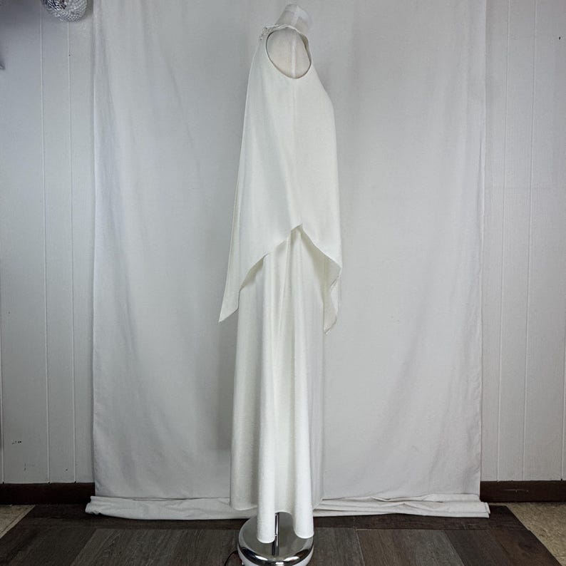 May include: A white, floor-length dress with a cape overlay. The dress has a halter neckline and a flowing, layered design. The fabric appears to be a lightweight material, creating a soft, elegant silhouette. The dress is displayed on a mannequin.