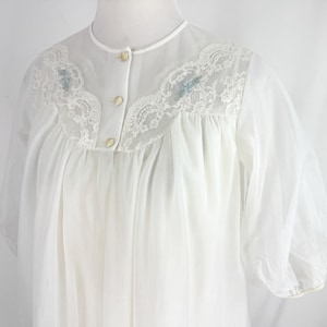 May include: A white vintage nightgown with a sheer overlay. The nightgown features a delicate lace yoke with embroidered floral accents in light blue. It has a button-down front and short, puffy sleeves.