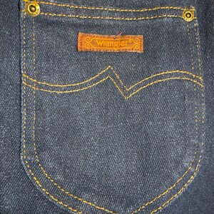 May include: Close-up of a dark blue denim pocket with gold stitching and brass rivets. A brown leather patch with the brand name "Wrangler" is centered on the pocket.