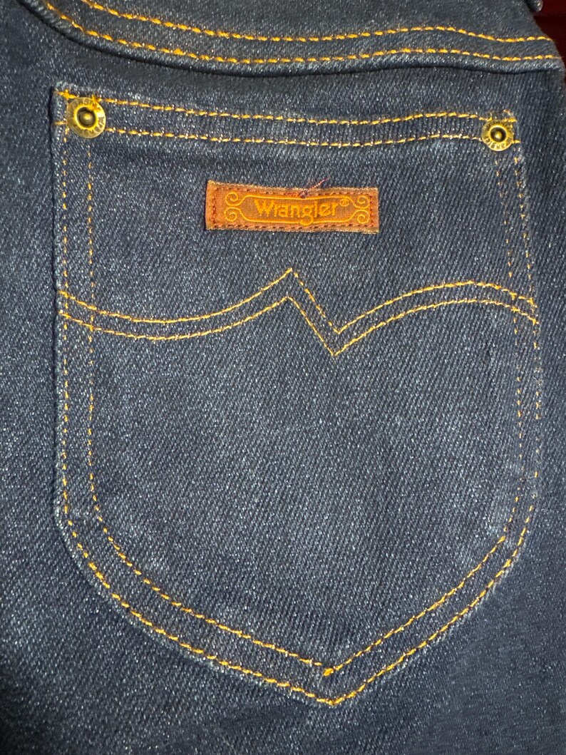 May include: Close-up of a dark blue denim pocket with gold stitching and brass rivets. A brown leather patch with the brand name "Wrangler" is centered on the pocket.