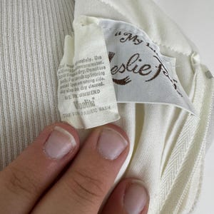 May include: Close-up of a cream-colored garment with a tag. The tag reads "My Leslie" in brown script, with care instructions and "Voolite THE FIR FABRIC WASH" printed below. The fabric has a ribbed texture.