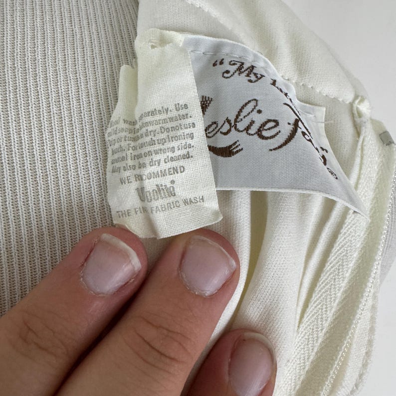 May include: Close-up of a cream-colored garment with a tag. The tag reads "My Leslie" in brown script, with care instructions and "Voolite THE FIR FABRIC WASH" printed below. The fabric has a ribbed texture.