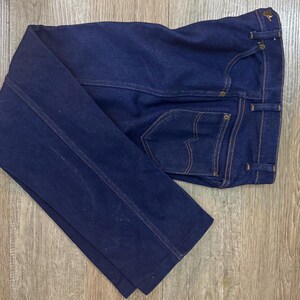 May include: Dark blue denim jeans with a classic five-pocket design. The jeans feature a straight leg cut, with visible stitching in a contrasting tan color. The back pockets have a distinctive curved design. The jeans are folded on a wooden surface.