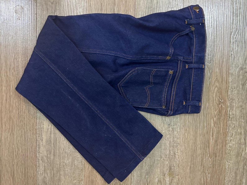 May include: Dark blue denim jeans with a classic five-pocket design. The jeans feature a straight leg cut, with visible stitching in a contrasting tan color. The back pockets have a distinctive curved design. The jeans are folded on a wooden surface.