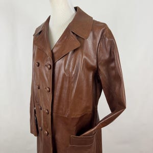 May include: A brown leather trench coat with a double breasted closure and a long length. The coat has a classic style and features a collar and pockets.