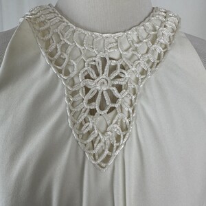 May include: Ivory-colored dress with a detailed, openwork crochet collar. The collar features a floral design at the center and a scalloped edge. The dress has a simple, elegant design.
