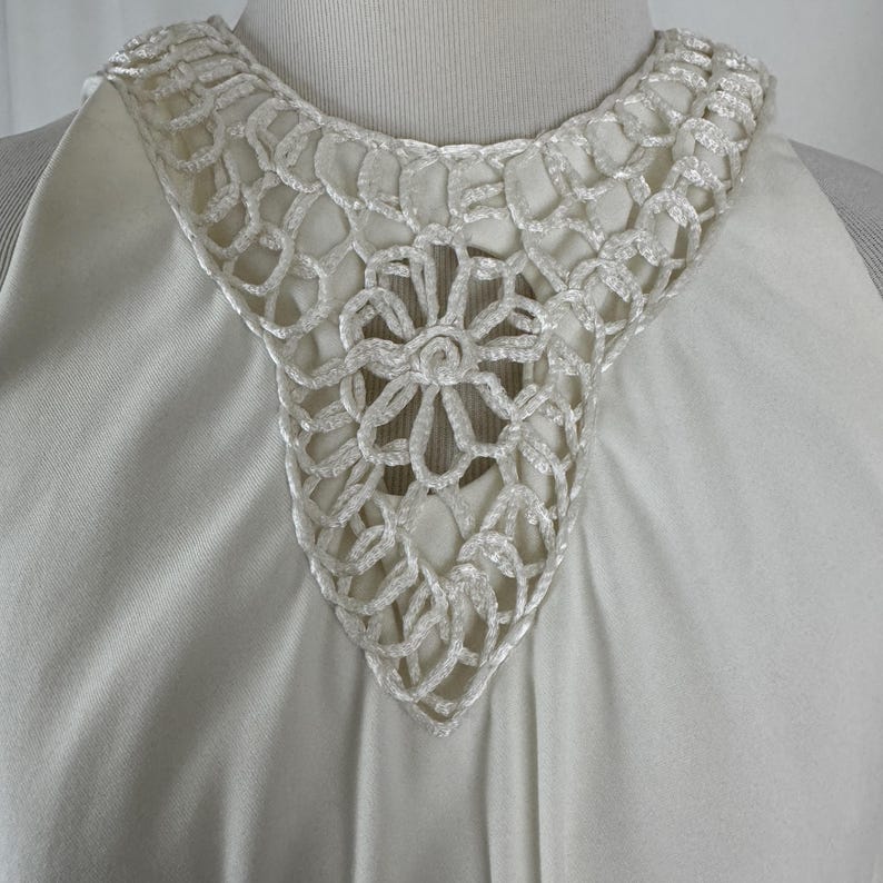 May include: Ivory-colored dress with a detailed, openwork crochet collar. The collar features a floral design at the center and a scalloped edge. The dress has a simple, elegant design.