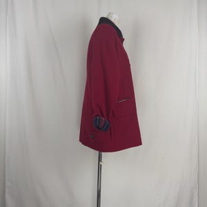 Op de afbeelding: Een bordeauxrode jas met een zwarte kraag en geruite manchetten. De jas heeft twee voorzakken en een ritssluiting op de borst. De jas wordt op een mannequin getoond.